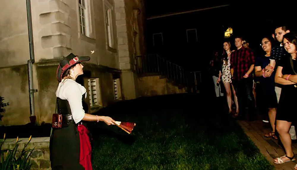 A person dressed in a pirate-inspired costume is leading a nighttime outdoor tour for a group of attentive onlookers