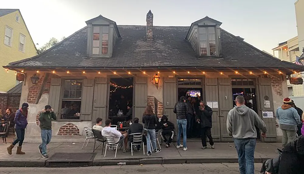 The image depicts a group of people socializing outside a charming rustic-looking bar adorned with string lights in the evening