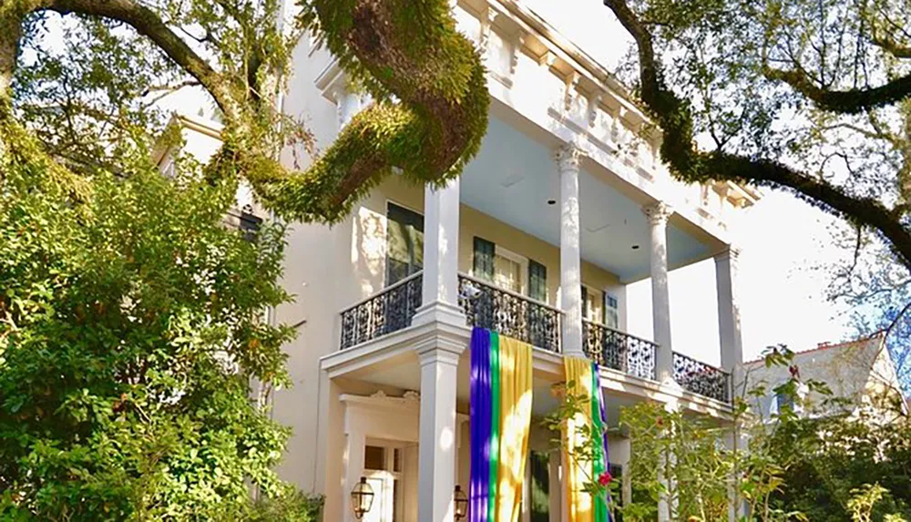 The image shows a grand white house with two tiers of columns and balconies adorned with Mardi Gras decorations nestled among lush greenery
