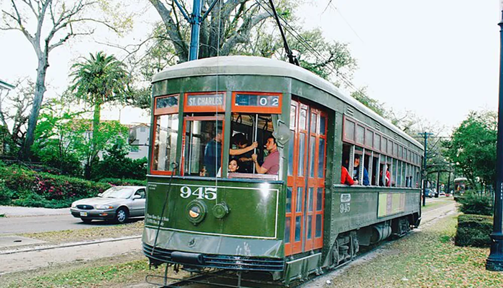 A green vintage streetcar labeled St Charles carrying passengers travels down a track-lined street flanked by trees and a car