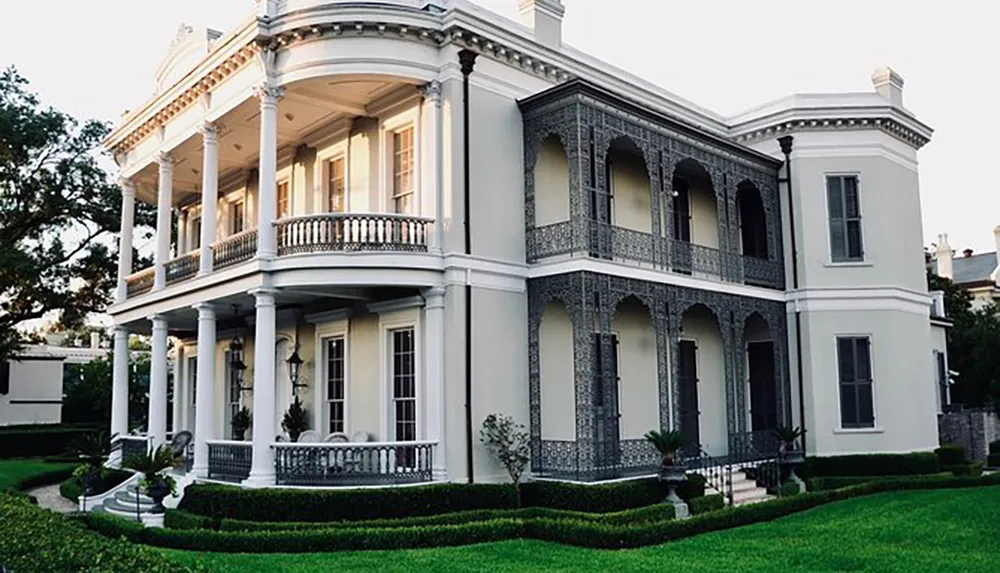 The image shows an elegant two-story mansion with white columns intricate ironwork on the balconies and manicured lawns suggesting an affluent Southern or historic estate