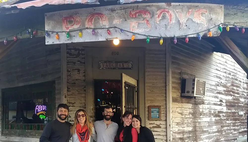 A group of friends are smiling for a photo in front of a rustic building with a sign reading PO-BOYS and colorful light bulbs strung overhead