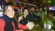 A group of friends is smiling and holding drinks at a bar decorated with festive lights.