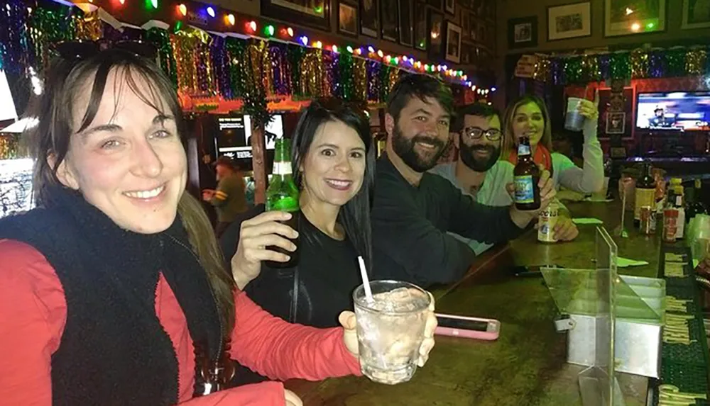 A group of friends is smiling and holding drinks at a bar decorated with festive lights