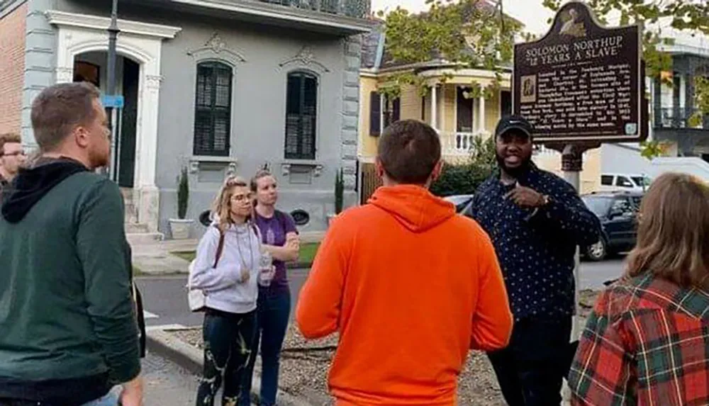 A group of people is attentively listening to a guide who is gesturing towards a historical marker about Solomon Northup near a street corner
