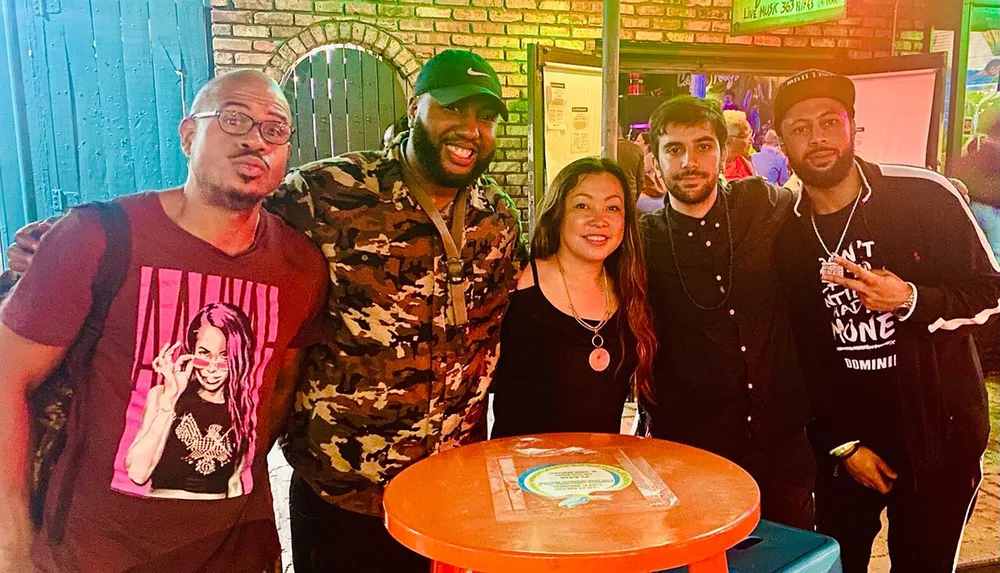 A group of five smiling people pose for a photo together at a venue with colorful lighting and a sign indicating live music is available 365 nights a year