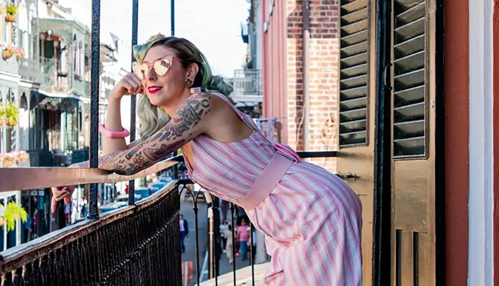 A woman with tattoos leans on a balcony railing smiling as she looks out onto a sunny street lined with historic-style buildings