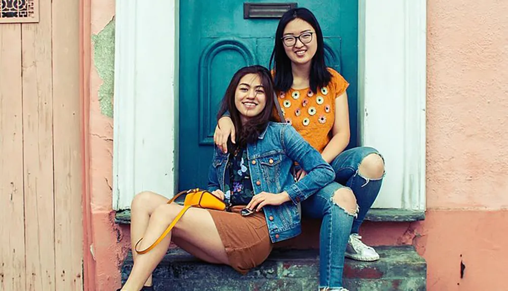 Two smiling women are sitting on a colorful doorstep exuding a sense of friendship and casual style
