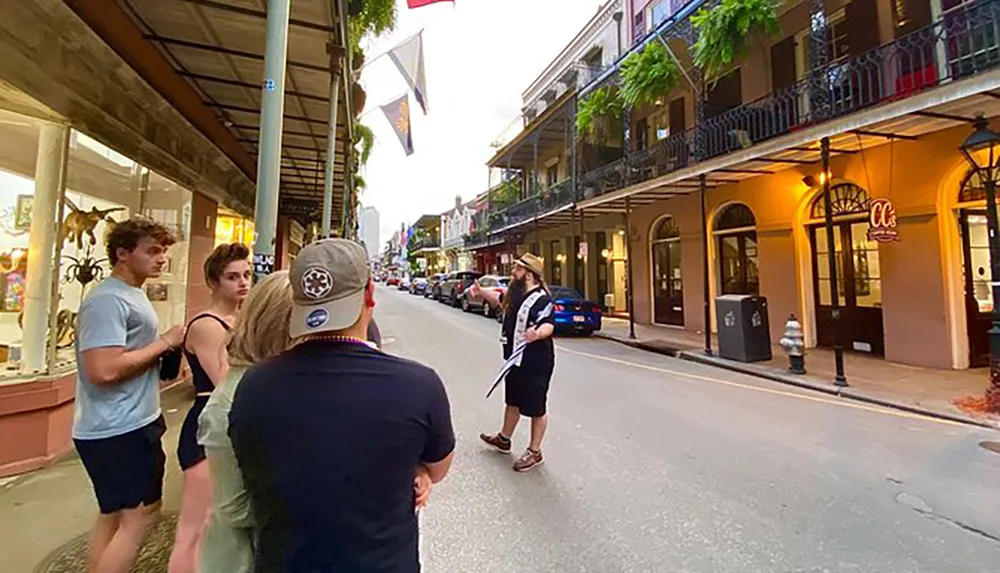 A group of people is walking down a street lined with traditional wrought-iron balconies and street lamps in what appears to be a historic urban area with a man in a wide-brimmed hat standing to one side