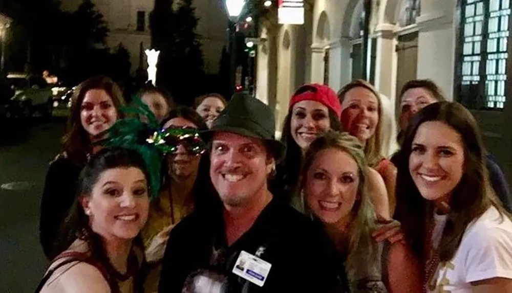 A group of joyous people mostly women wearing various accessories such as hats and masks with one man in the center wearing a hat all smiling for a group photo at night on a city street