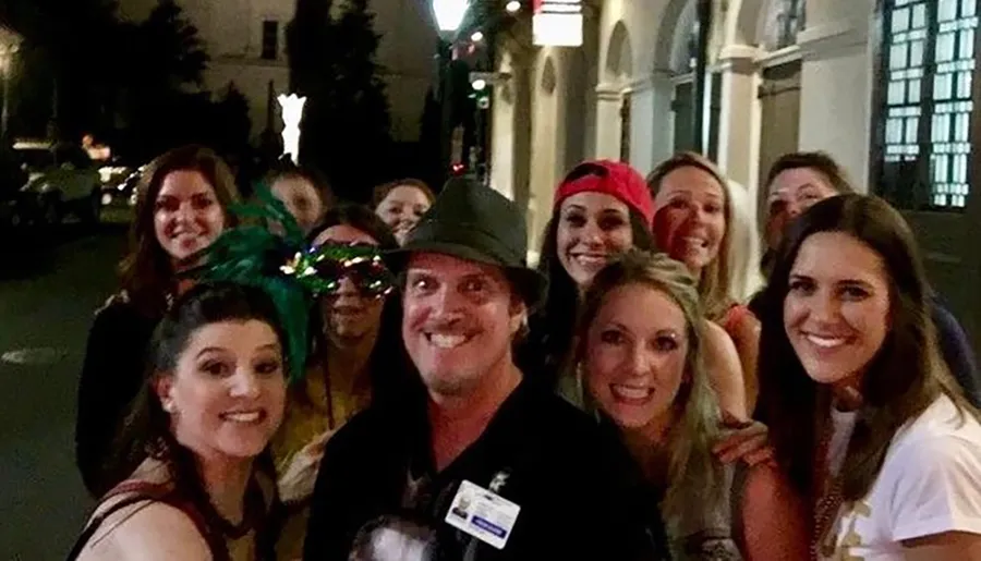 A group of joyous people, mostly women wearing various accessories such as hats and masks, with one man in the center wearing a hat, all smiling for a group photo at night on a city street.
