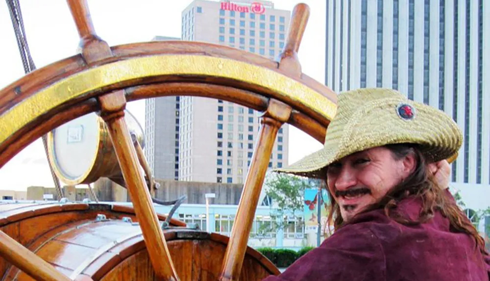 A person with a goatee and wearing a straw hat is smiling at the camera while standing behind a large wooden ships wheel with urban high-rise buildings including one with a Hilton hotel logo in the background