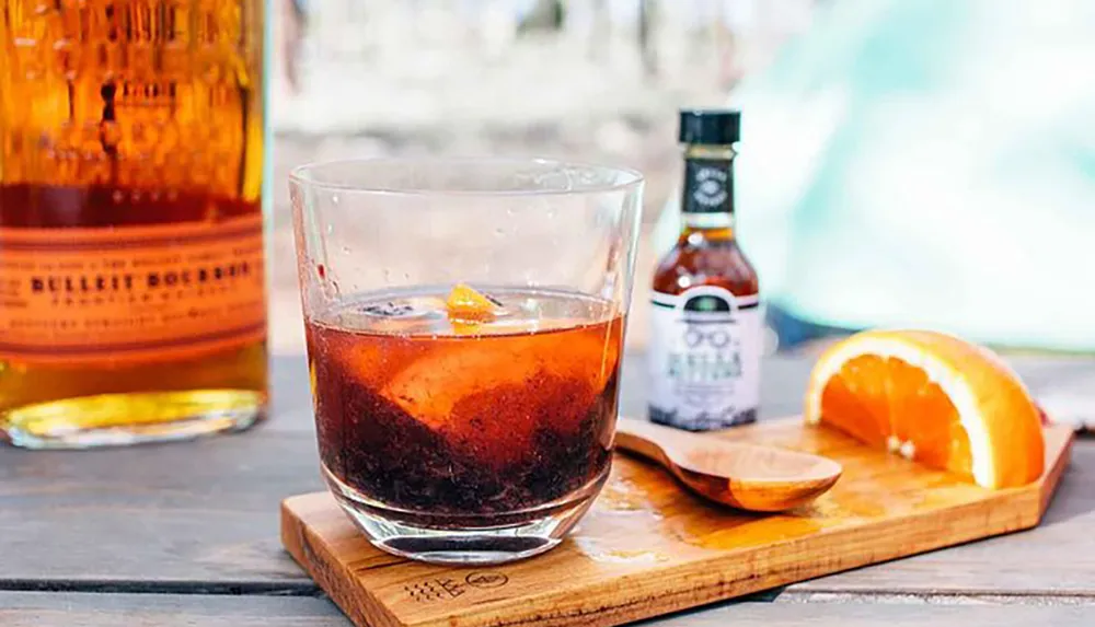 A glass of bourbon cocktail with ice and a twist of orange peel accompanied by a bottle of bourbon and a sliced orange on a wooden board
