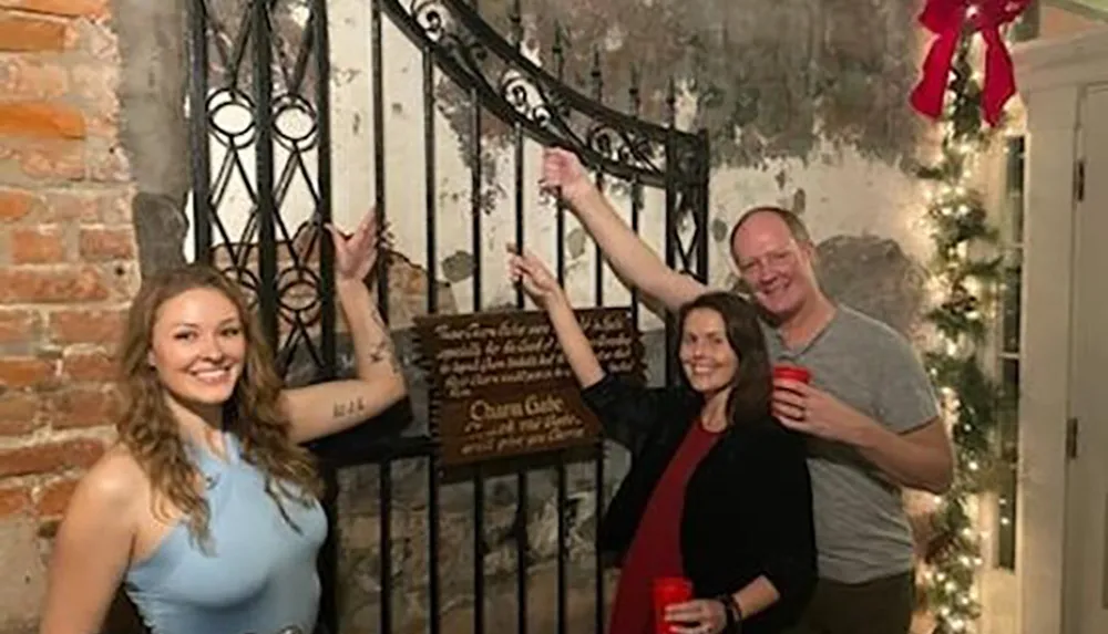 Three people are cheerfully posing before an ornamental iron gate and a sign in a room with exposed brick walls and holiday decorations