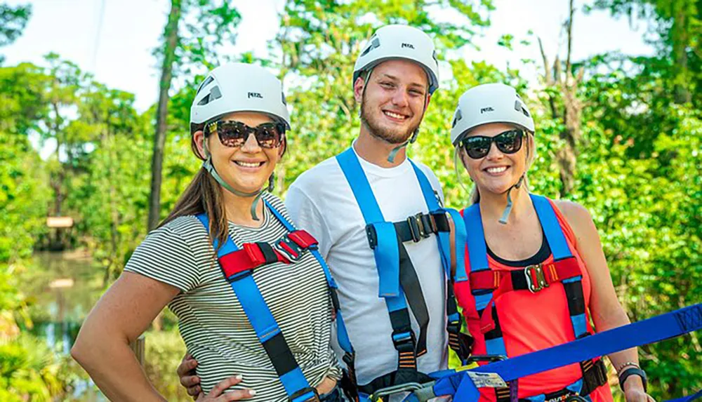 Three people are smiling outdoors wearing safety helmets and harnesses possibly preparing for an adventure activity such as zip-lining or rock climbing