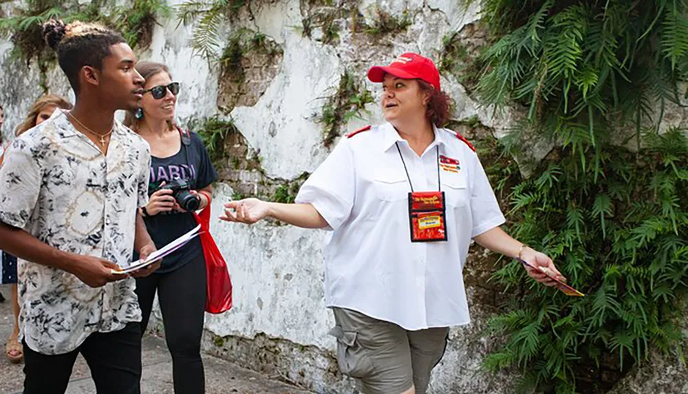 A tour guide in a red hat and white shirt is addressing tourists one holding a camera and another a booklet against a backdrop with lush greenery