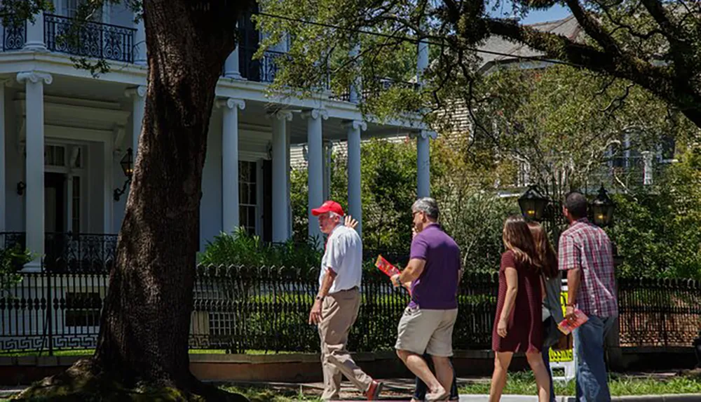 A group of people are walking and talking near a white-pillared house shaded by large trees on a sunny day