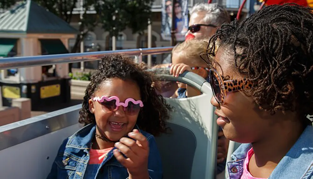 A child with wavy sunglasses is laughing joyfully while riding on a tour bus next to an adult