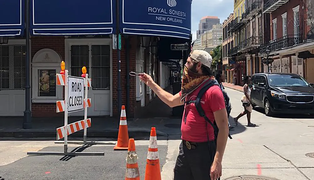 A person is looking at their phone on a sunny street next to a ROAD CLOSED sign with orange traffic cones and the Royal Sonesta New Orleans canopy in the background