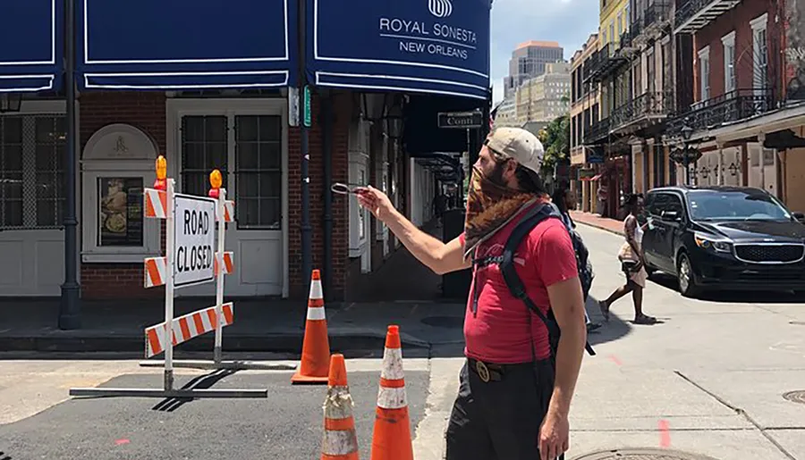 A person is looking at their phone on a sunny street next to a ROAD CLOSED sign with orange traffic cones and the Royal Sonesta New Orleans canopy in the background.