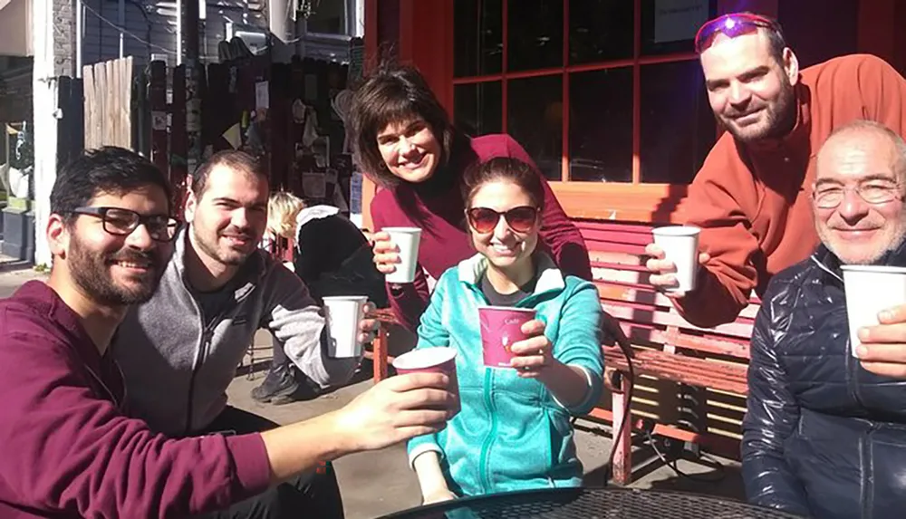 A group of five people are gathered around a table outside smiling and toasting with their coffee cups in a casual sunny setting
