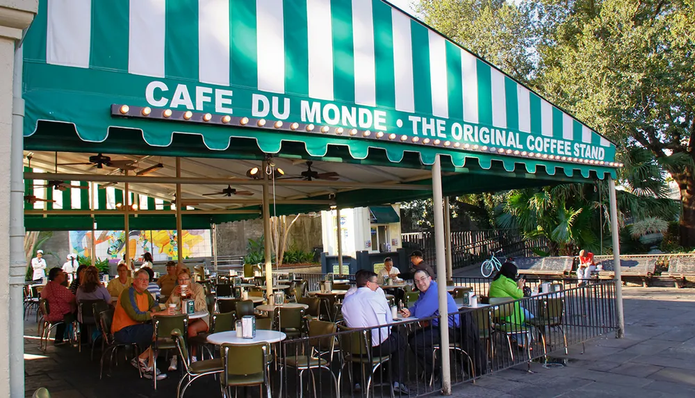 The image shows people seated at outdoor tables under a green and white striped canopy with the sign CAFE DU MONDE  THE ORIGINAL COFFEE STAND in a casual social atmosphere