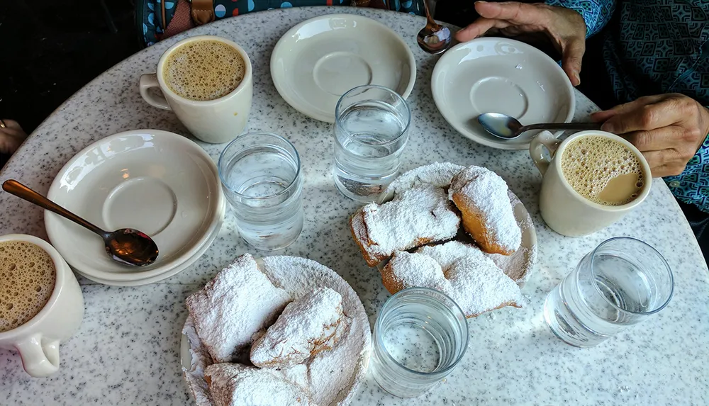 A table is seen with hands of two people cups of coffee plates glasses of water and beignets covered in powdered sugar