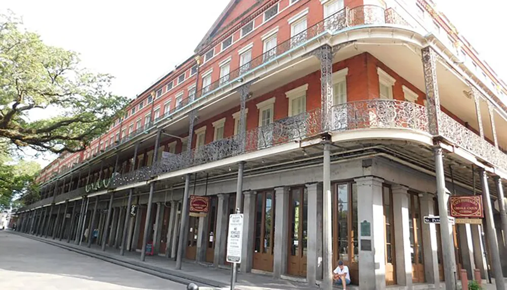 The image shows a three-story red building with ornate iron balconies ground-floor columns and a person sitting on the sidewalk which is likely located in a historic district reminiscent of New Orleans French Quarter