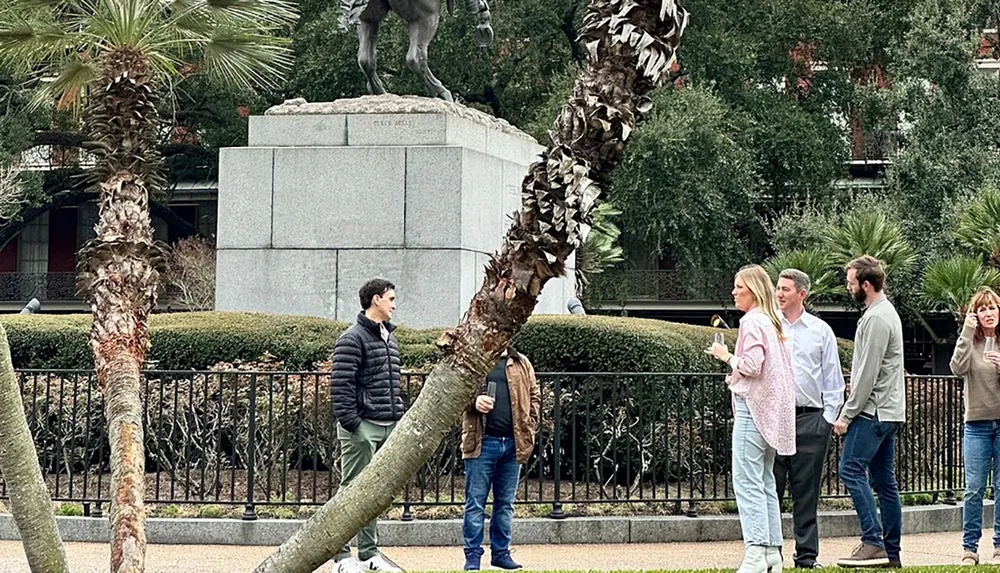 The image shows a group of people casually conversing in an outdoor setting with palm trees and a statue in the background