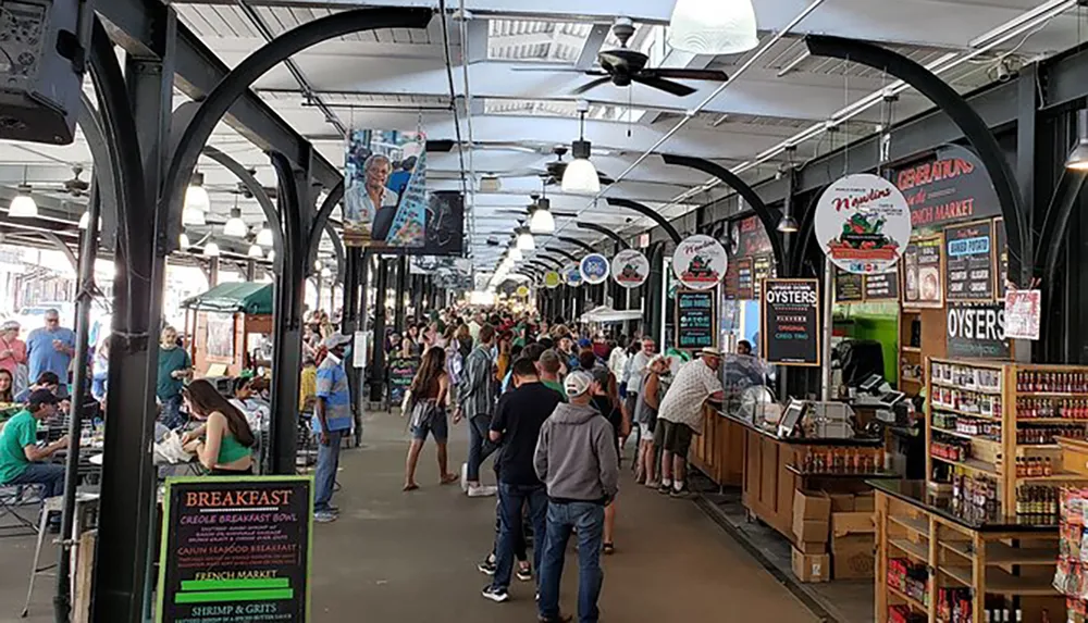 The image displays a bustling indoor market with people browsing various food stalls and seating areas under an arched metal ceiling structure