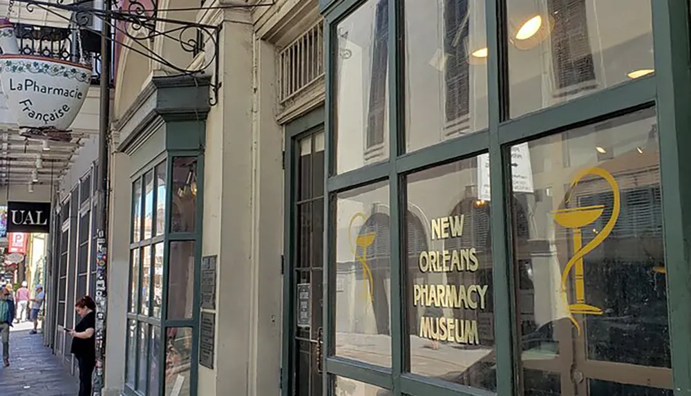 The image shows the exterior of the New Orleans Pharmacy Museum with signage and a caduceus symbol on its window indicating its historical and educational significance