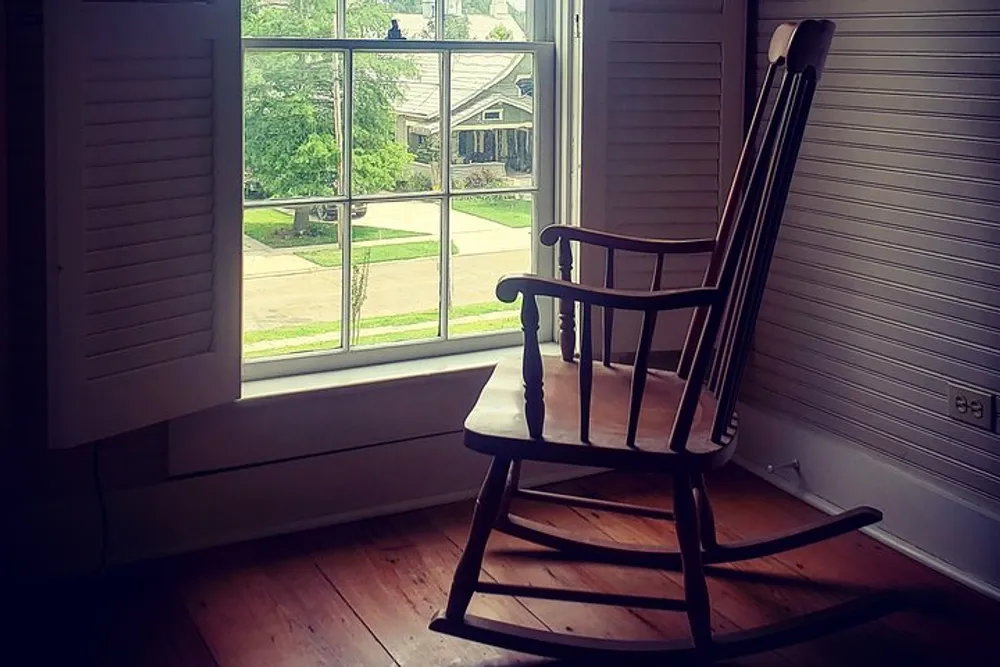A wooden rocking chair is situated near a window with open shutters overlooking a street view outside a residential home