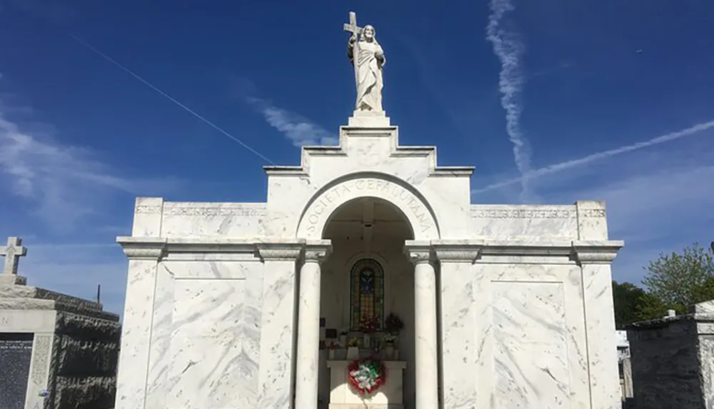 An ornate white marble mausoleum with a statue of Jesus holding a cross stands under a clear blue sky with streaks of airplane contrails