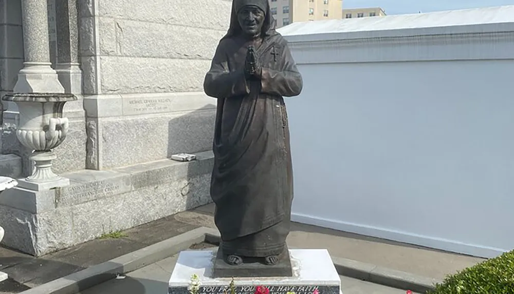 This image shows a dark-toned statue of a person standing with hands clasped in prayer mounted on a white base with text and located beside a building and bush