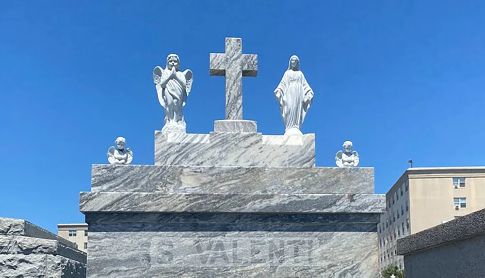 The image shows a marble religious monument featuring statues of angels a cross and the Virgin Mary against a clear blue sky
