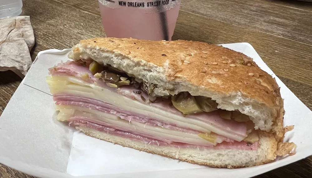 A half-eaten muffuletta sandwich rests on a plate showcasing layers of meat cheese and olive salad with a pink beverage in the background