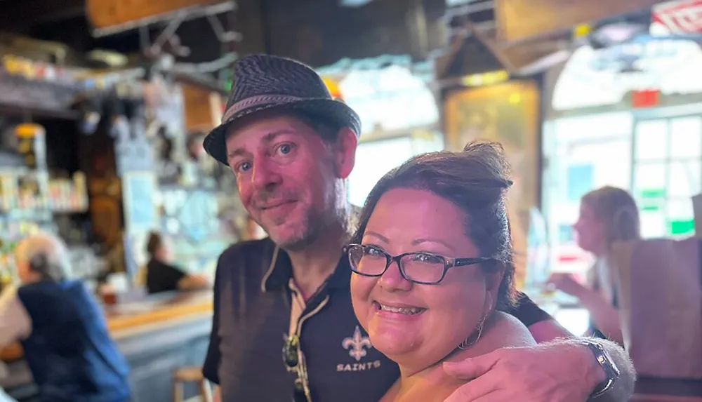 A man wearing a hat and a woman with glasses are smiling together for a photo in a bar with a blurred background