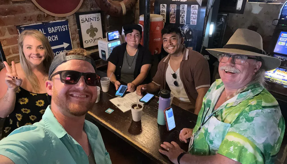 A group of five people is smiling for a selfie inside a bar with some holding drinks and phones on the bar top