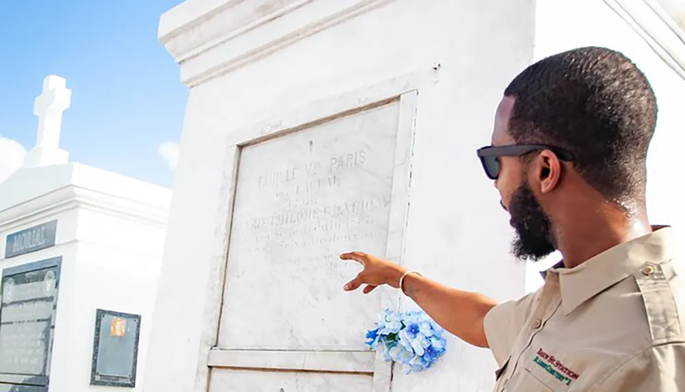 A man in sunglasses and a light-colored shirt is gesturing towards an inscription on a white mausoleum adorned with a blue floral tribute under a bright sky