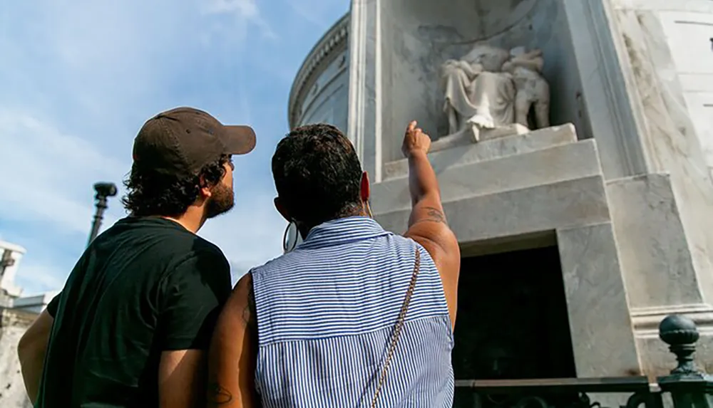 Two people are standing with their backs to the camera looking up at a neoclassical monument with an angel statue under a clear sky