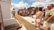 A group of tourists is attentively listening to a guide who is gesturing as he speaks, amidst the historical setting of an old cemetery on a sunny day.