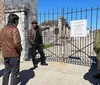 A group of people are engaged in what appears to be a guided tour outside the gates of the St Louis Cemetery 2 with the guide gesturing towards something inside the cemetery