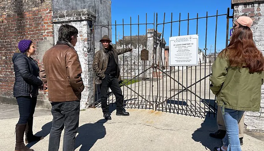 A group of people are engaged in what appears to be a guided tour outside the gates of the St. Louis Cemetery #2, with the guide gesturing towards something inside the cemetery.