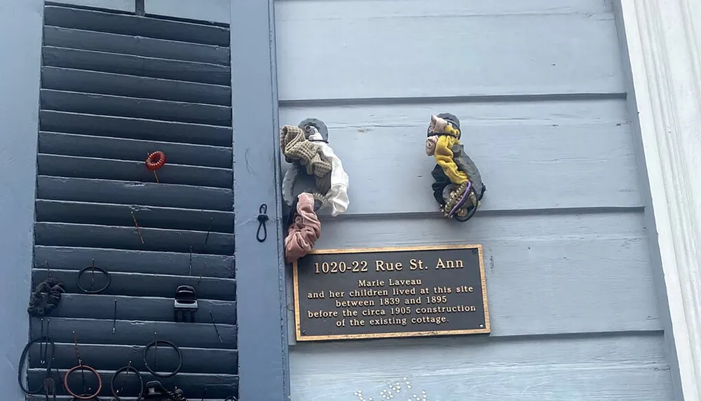 The image shows a plaque on a wall commemorating Marie Laveaus residence at this site flanked by two sculptural elements with a collection of metal rings and objects mounted below on the shutter