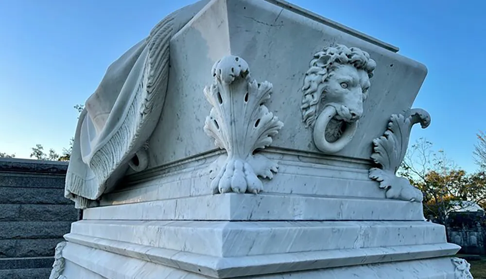 The image shows a white marble sculpture with a stylized lions head and classical decorative elements including acanthus leaves and architectural flourishes
