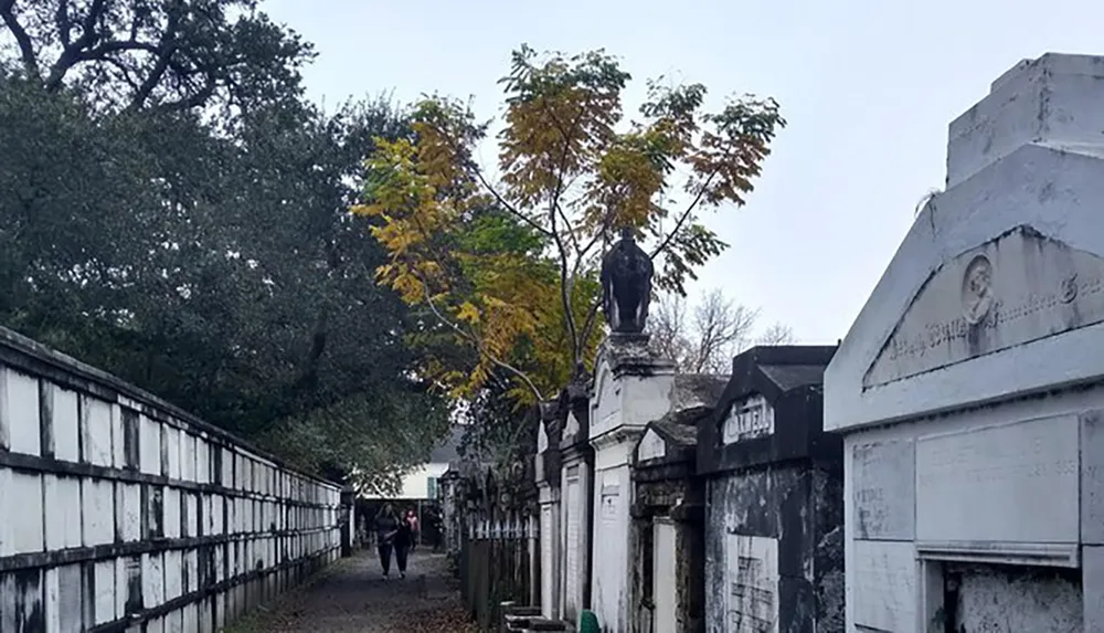 Two people stroll past the weathered tombs and mausoleums lined along a pathway in an atmospheric cemetery