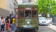 Passengers board an olive-green St. Charles streetcar in a city setting, surrounded by cars and pedestrians.