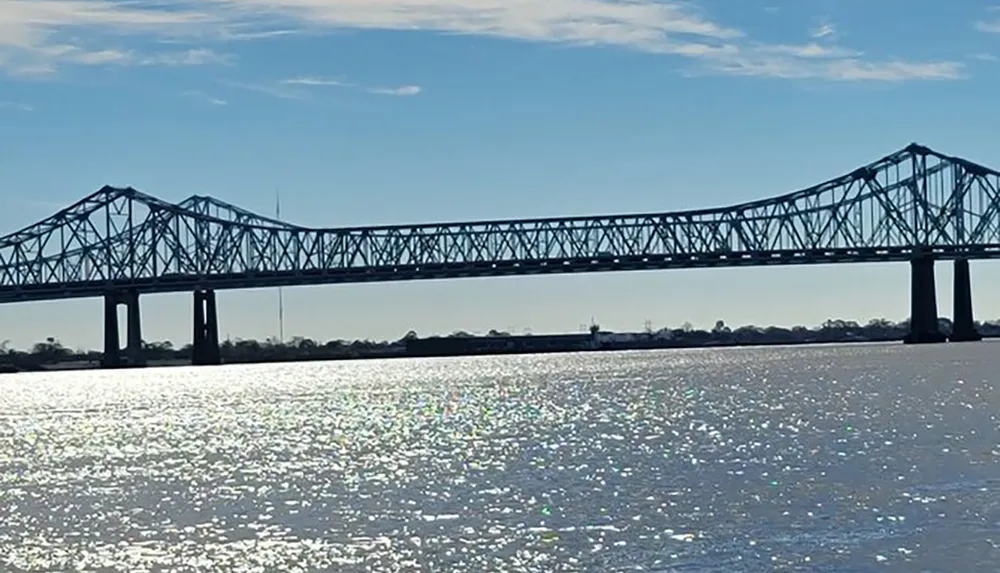 The image shows a large metal truss bridge spanning over a shimmering body of water under a clear sky
