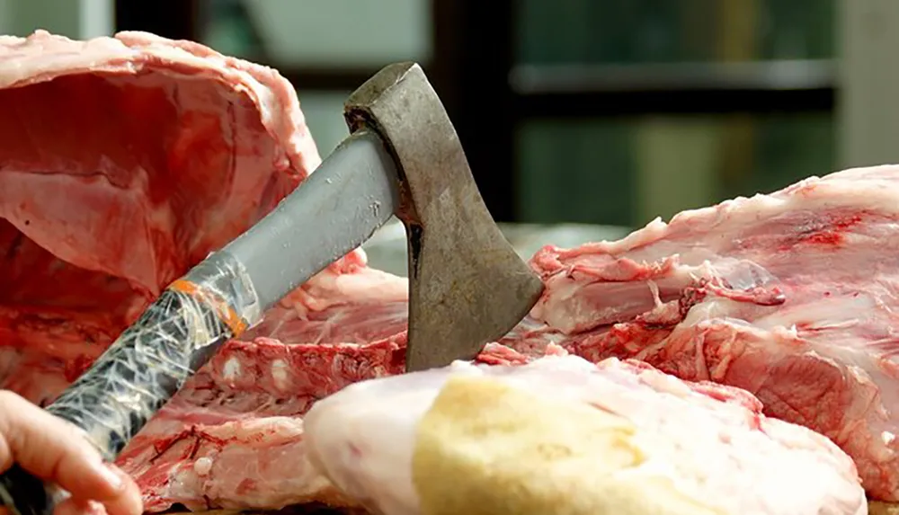A persons hand is holding a cleaver embedded in a large piece of raw meat on a cutting surface