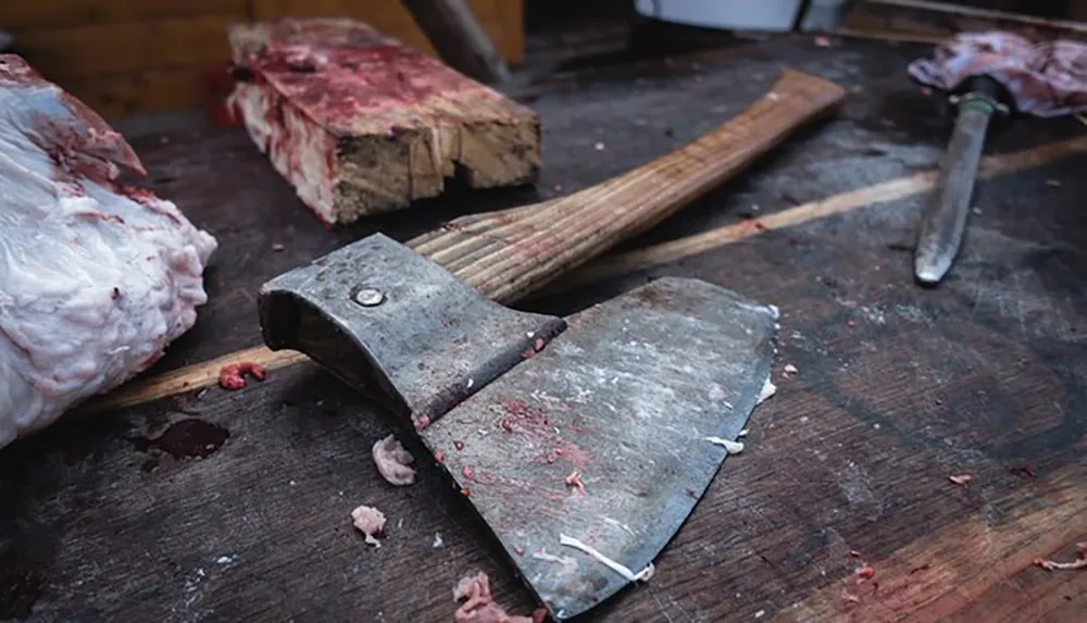 A cleaver and a knife rest on a worn wooden cutting board beside chunks of raw meat